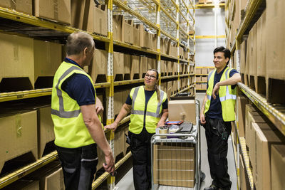 Multi-ethnic coworkers discussing while standing with cart on aisle amidst racks at distribution warehouse