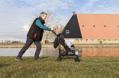 Grandfather pushing granddaughter while sitting in baby stroller by canal in city