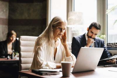 Mature businesswoman wearing in-ear headphones looking at laptop by businessman in office