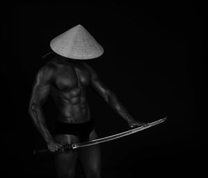 Muscular man in hat with sword standing against black background