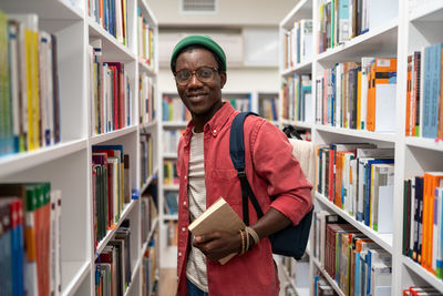 Smiling african american man student holds book in university library between bookshelves.