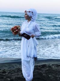 Beautiful young bride standing on shore at beach against sky during sunset