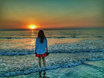 Rear view of woman standing at sea shore against sky during sunset