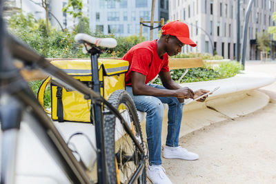 Young delivery man using digital tablet while sitting by package on bench