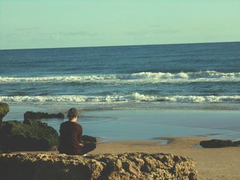 Rear view of woman sitting on rock by sea against sky
