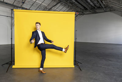 Businessman standing on one leg raising foot in front of yellow backdrop