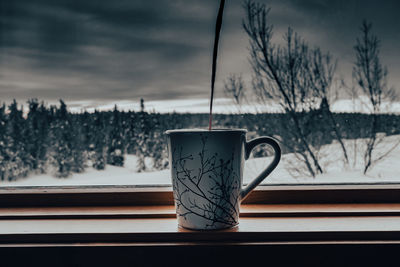 Close-up of coffee cup against bare trees during winter