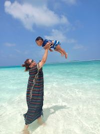 Woman tossing toddler son while standing in sea against blue sky