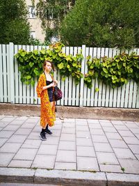 Full length portrait of girl standing on footpath against fence