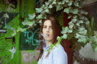 Portrait of beautiful young woman emitting smoke from mouth while standing against wall