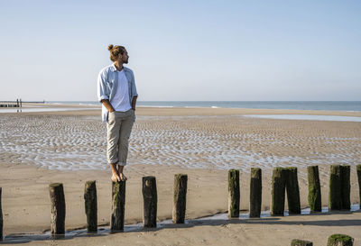 Young man looking at view while standing on wooden post at beach against clear sky