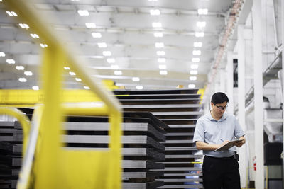 Mature supervisor writing on clipboard while standing by metal sheets at warehouse