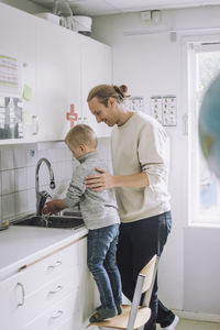 Male teacher guiding boy to wash hands standing on chair near sink at kindergarten