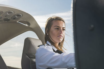 Low angle view of female pilot operating control panel while sitting in airplane against blue sky at airport