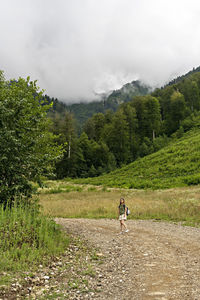 Woman on dirt road amidst trees and mountains against sky