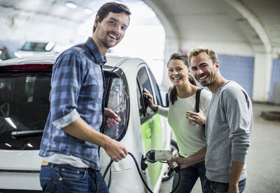 Portrait of happy friends with man charging electric car at gas station