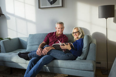 Smiling couple reading book while resting on sofa in living room at home