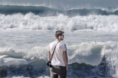 Rear view of man carrying camera standing against splashing sea waves