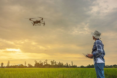 A farmer in a cap stands in a lush wheat field, directing a drone that is flying above the