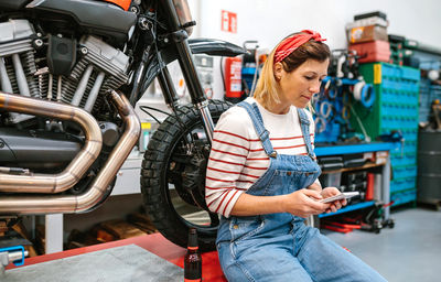 Mechanic woman looking phone sitting over platform with motorcycle on factory