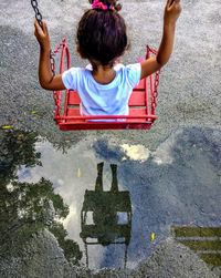 Rear view of girl swinging reflecting on puddle during rainy season