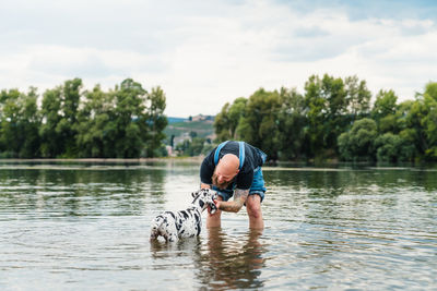 Happy man with beard in casual clothes playing with puppy while standing in lake water in summer