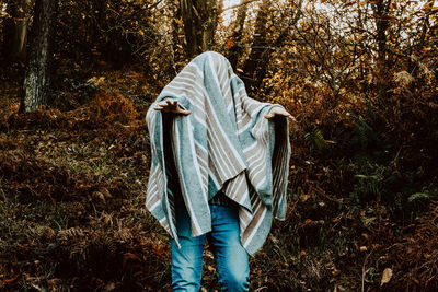 Man covered in blanket gesturing while standing in forest 
