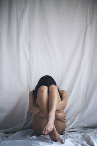 Full length of naked young woman sitting on bed against textile