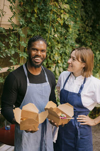 Smiling female chef with hand on hip looking at male owner holding food boxes at restaurant