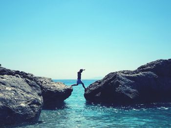 Man on rock by sea against clear blue sky