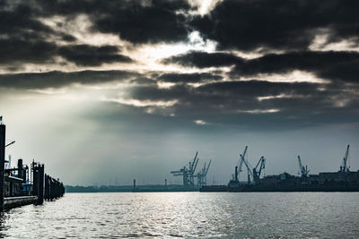 Silhouette cranes at harbor against sky