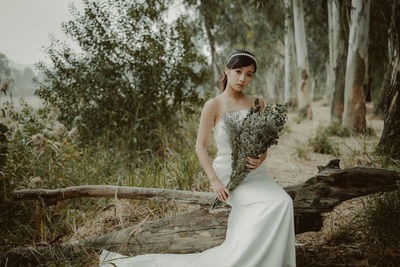 Portrait of woman holding bouquet while sitting on log in forest