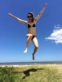 Full length of woman in bikini jumping on grassy field at beach against blue sky during sunny day