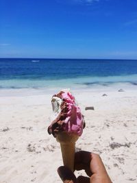 Cropped image of hand holding ice cream at beach against blue sky