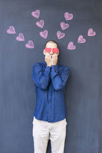 Man with heart shape paper on eyes standing against blackboard