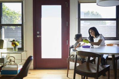 Mother feeding water to son from bottle while sitting against window at home