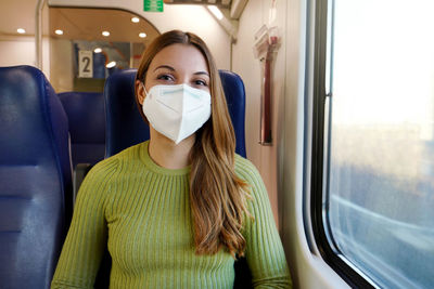 Happy young woman wearing medical mask sitting on train looking at camera.