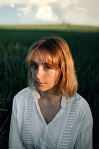 Peaceful young female in retro styled white blouse sitting amidst tall green grass and looking at camera while resting in summer evening in countryside