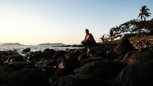 Shirtless man crouching on rocky shore against clear sky