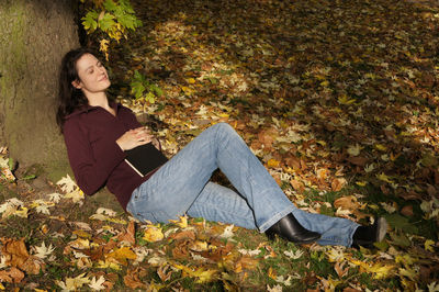 Smiling woman with book reclining on tree at park during autumn