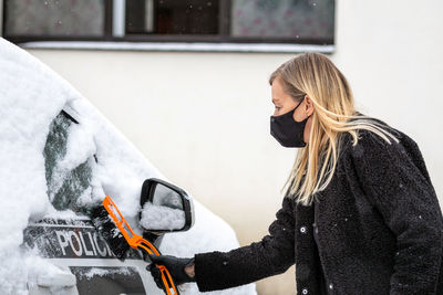 Blond woman in face mask with a squeegee cleans snow from a police car