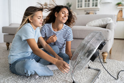 Mother and daughter cooling down while sitting on floor in front of electric fan blowing fresh air