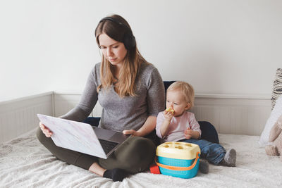 Mother using laptop while sitting with baby girl on bed at home