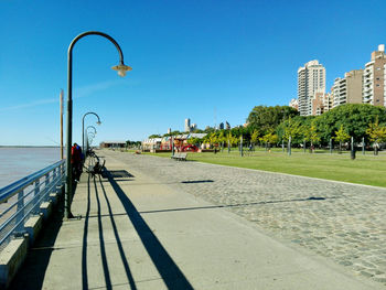 Railing by sidewalk with trees and buildings against blue sky