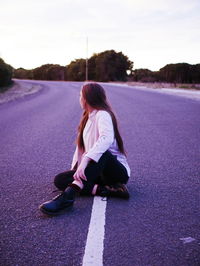 Young woman sitting on road against sky at sunset