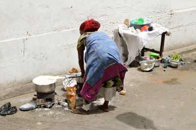 Rear view of poor woman cooking on stove by wall on street