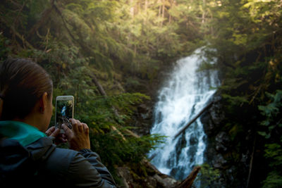 Rear view of woman photographing waterfall at forest