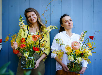 Two young women with beautiful bouquets of peonies in glass vases against the blue background