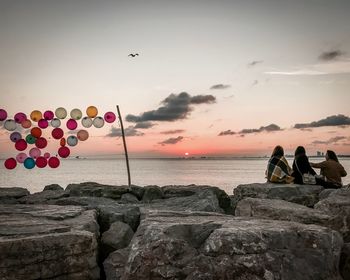 People on rocks by sea against sky during sunset