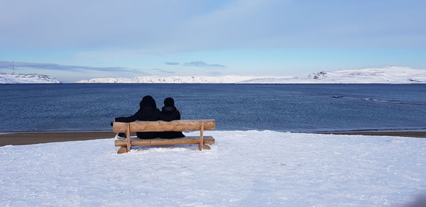 Rear view of friends sitting on bench by lake against sky during winter
